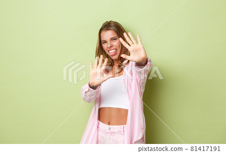 Image of displeased young blond woman defending herself from bright light, raising hands and grimacing bothered, asking to stop taking pictures of her, standing over green background Image of displeased young blond woman defending herself from bright light, raising hands and grimacing bothered, asking to stop taking pictures of her, standing over green background 81417191