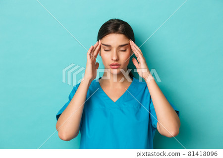 Coronavirus, social distancing and health concept. Close-up of woman doctor in scrubs feeling exhausted, having a headache, standing over blue background 81418096