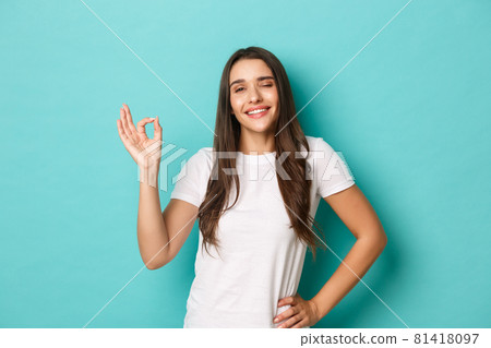 Image of confident smiling woman in white t-shirt, showing okay sign and looking pleased, agree or approve something good, like and recommend product, standing over blue background 81418097