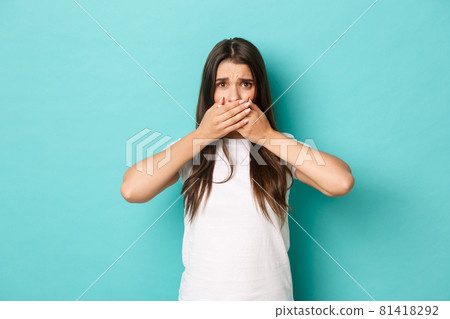 Image of scared brunette girl in white t-shirt, looking anxious and shut mouth with hands, standing over blue background 81418292