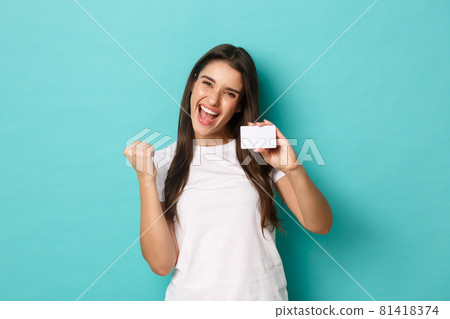 Image of satisfied ecstatic woman, wearing white t-shirt, rejoicing and showing credit card, standing pleased over blue background Image of satisfied ecstatic woman, wearing white t-shirt, rejoicing and showing credit card, standing pleased over blue background 81418374
