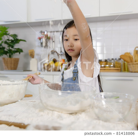 A little cute Asian girl, 7 years old, learning how to make bread and bakery, she plays and tries to mix flour with a curious and happy smiley face in a modern kitchen. A little cute Asian girl, 7 years old, learning how to make bread and bakery, she plays and tries to mix flour with a curious and happy smiley face in a modern kitchen. 81418806