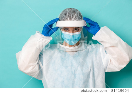 Concept of covid-19, pandemic and quarantine. Close-up of serious female doctor put on face shield and personal protective equipment to enter contagious zone, standing over blue background 81419604