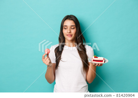 Image of young slim girl in white t-shirt, tempted to eat macaroon, holding cake, standing over blue background Image of young slim girl in white t-shirt, tempted to eat macaroon, holding cake, standing over blue background 81420904
