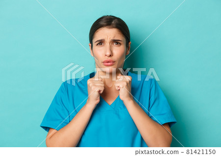 Coronavirus, social distancing and health concept. Close-up of worried young nurse in scrubs, looking concerned at something bad, grimacing disturbed, standing over blue background 81421150