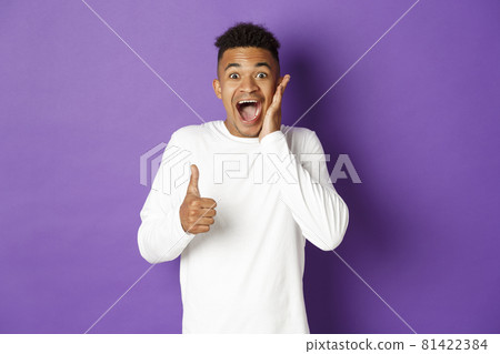 Portrait of amazed african-american man, showing thumbs-up and looking excited, recommend and praise good product, standing over purple background 81422384