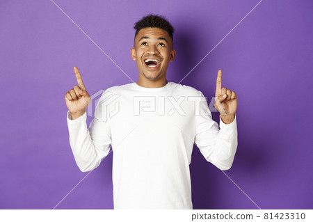 Handsome excited african-american guy, wearing white sweatshirt, looking amazed and pointing fingers up, showing super cool advertisement, standing over purple background Handsome excited african-american guy, wearing white sweatshirt, looking amazed and pointing fingers up, showing super cool advertisement, standing over purple background 81423310