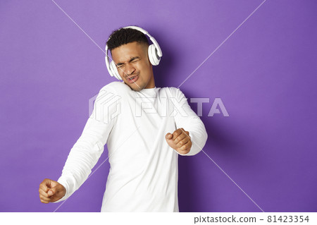 Image of carefree african-american guy dancing, listening music in wireless headphones, standing over purple background 81423354