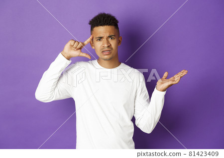 Image of angry african-american man, looking confused, pointing at head and scolding person for stupid action, standing over purple background 81423409
