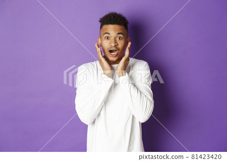 Portrait of handsome african-american male model in white sweatshirt, looking amazed and impressed, gasping wondered, standing over purple background Portrait of handsome african-american male model in white sweatshirt, looking amazed and impressed, gasping wondered, standing over purple background 81423420