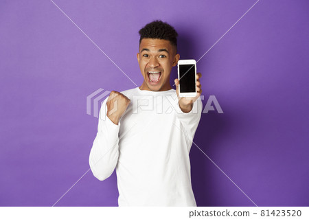 Excited african-american man showing mobile phone screen, winning something online, rejoicing over app achievement, standing over purple background 81423520