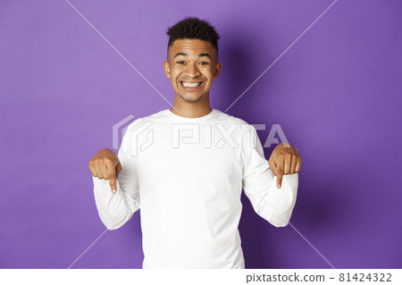 Portrait of african-american happy guy, wearing white sweatshirt, pointing fingers down and smiling cheerful, showing advertisement, standing over purple background 81424322