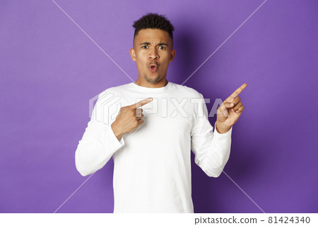 Portrait of confused and shocked african-american guy in white sweatshirt, pointing fingers right and frowning, showing something strange, standing over purple background 81424340