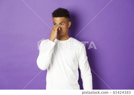 Portrait of young african-american guy in white sweatshirt, shut nose and grimacing from bad smell, looking at something nasty with disgusting reek, standing over purple background 81424383