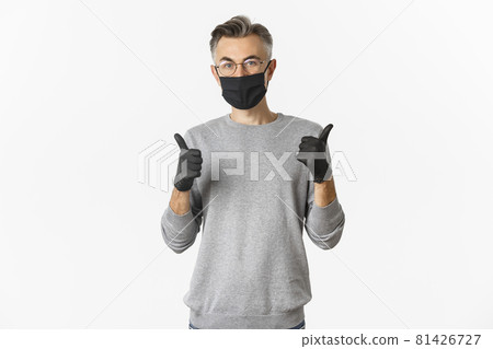 Covid-19, pandemic and social distancing concept. Image of middle-aged man in black medical mask, gloves and glasses showing thumbs-up, protect himself from catching coronavirus Covid-19, pandemic and social distancing concept. Image of middle-aged man in black medical mask, gloves and glasses showing thumbs-up, protect himself from catching coronavirus 81426727