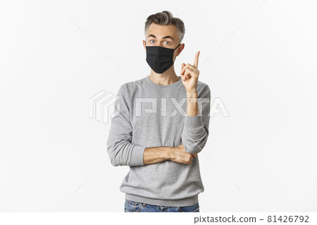 Covid-19, pandemic and social distancing concept. Portrait of handsome middle-aged caucasian man, wearing medical mask, having suggestion or idea, raising finger in eureka sign, white background Covid-19, pandemic and social distancing concept. Portrait of handsome middle-aged caucasian man, wearing medical mask, having suggestion or idea, raising finger in eureka sign, white background 81426792