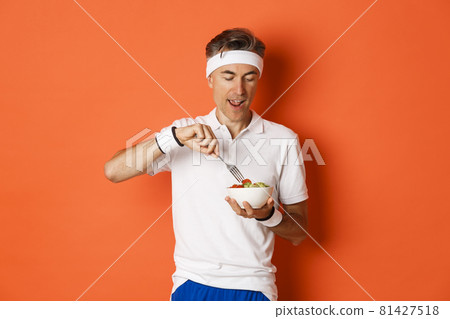 Concept of sport, fitness and lifestyle. Portrait of joyful middle-aged guy in workout uniform, holding fork and salad, eating healthy food, standing over orange background 81427518