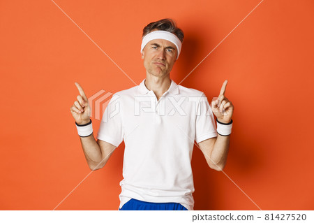 Portrait of skeptical adult sportsman in headband and t-shirt, complaining and pointing fingers up, looking disappointed, standing over orange background Portrait of skeptical adult sportsman in headband and t-shirt, complaining and pointing fingers up, looking disappointed, standing over orange background 81427520