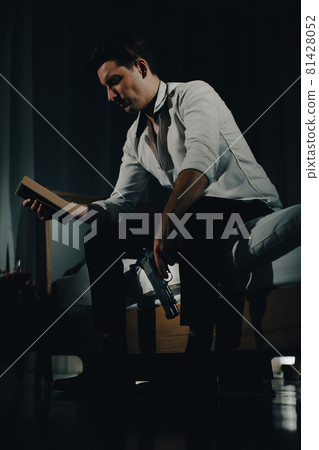 A young man in casual business clothes sitting alone in bedroom holding a pistol and looking at family photo in frame look like going to kill himself. Concept of hopelessness and give up people A young man in casual business clothes sitting alone in bedroom holding a pistol and looking at family photo in frame look like going to kill himself. Concept of hopelessness and give up people 81428052