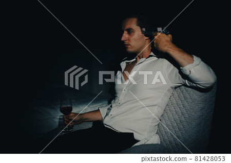 A young man in casual business clothes sitting alone in dark bedroom holding a pistol aim at his head and wine glass looks like going to kill himself. Concept of hopelessness and give up people A young man in casual business clothes sitting alone in dark bedroom holding a pistol aim at his head and wine glass looks like going to kill himself. Concept of hopelessness and give up people 81428053