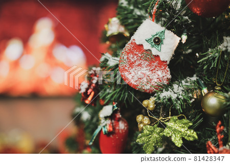 Closeup shot of decorated sock hanging on pine branch of Christmas tree with a ball and various decor xmas ornaments cover with snowflakes and blur background of fire flame in fireplace Closeup shot of decorated sock hanging on pine branch of Christmas tree with a ball and various decor xmas ornaments cover with snowflakes and blur background of fire flame in fireplace 81428347
