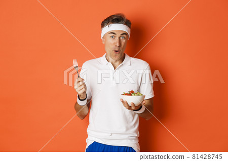 Concept of sport, fitness and lifestyle. Portrait of joyful middle-aged guy in workout uniform, holding fork and salad, eating healthy, standing over orange background Concept of sport, fitness and lifestyle. Portrait of joyful middle-aged guy in workout uniform, holding fork and salad, eating healthy, standing over orange background 81428745