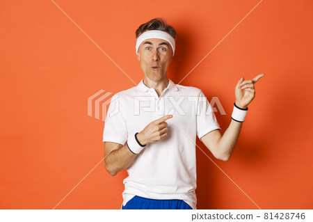 Concept of workout, sports and lifestyle. Portrait of amused middle-aged male athlete, pointing fingers at upper right corner, showing cool promo offer, standing over orange background 81428746