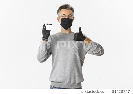 Concept of covid-19, social distancing and lifestyle. Satisfied middle-aged man in medical mask, gloves and glasses, recommending bank, showing credit card and thumbs-up Concept of covid-19, social distancing and lifestyle. Satisfied middle-aged man in medical mask, gloves and glasses, recommending bank, showing credit card and thumbs-up 81428747