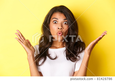 Close-up of surprised african-american girl, raising hands up and saying wow, hear amazing news, standing over yellow background Close-up of surprised african-american girl, raising hands up and saying wow, hear amazing news, standing over yellow background 81428786