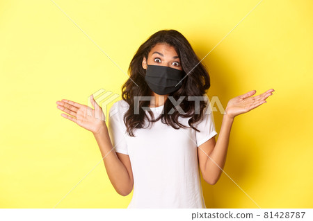 Concept of coronavirus, pandemic and lifestyle. Image of cute african-american girl in face mask, shrugging and looking clueless, dont know anything, standing over yellow background Concept of coronavirus, pandemic and lifestyle. Image of cute african-american girl in face mask, shrugging and looking clueless, dont know anything, standing over yellow background 81428787