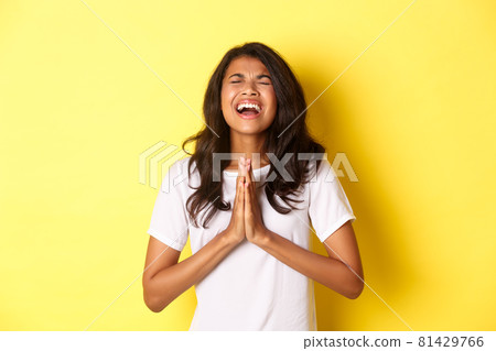 Portrait of young african-american girl pleading, begging for help with hands pressed together on chest, standing in white t-shirt over yellow background 81429766