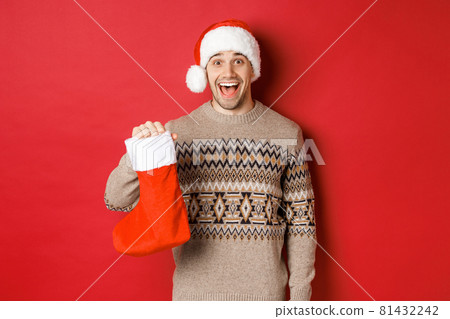 Concept of winter holidays, new year and celebration. Cheerful and surprised adult man receiving candies on Saint Nicholas day in red stocking, standing amazed over red background Concept of winter holidays, new year and celebration. Cheerful and surprised adult man receiving candies on Saint Nicholas day in red stocking, standing amazed over red background 81432242