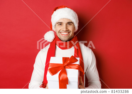 Concept of winter holidays, christmas and lifestyle. Close-up of attractive bearded guy in santa hat and scarf, holding a present, giving new year gift and smiling, red background 81433286