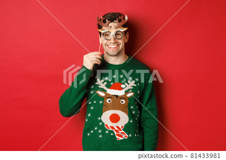 Handsome happy man in christmas sweater, holding party mask and smiling, enjoying new year celebration, standing over red background 81433981