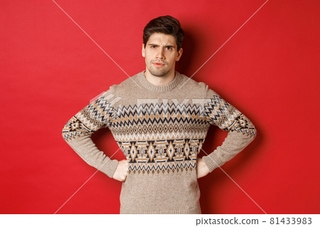 Portrait of doubtful and disappointed man, wearing christmas sweater, frowning and looking displeased at you, standing against red background 81433983