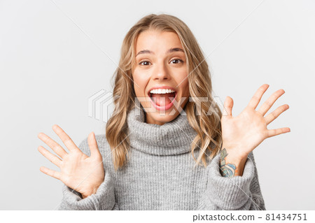 Close-up of friendly attractive girl in grey sweater, smiling and saying hi, waving hand to greet someone, standing over white background Close-up of friendly attractive girl in grey sweater, smiling and saying hi, waving hand to greet someone, standing over white background 81434751