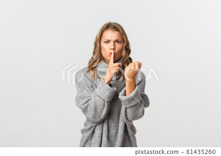 Close-up of grumpy and angry blond girl in grey sweater, scolding someone for being loud, shaking fist and shushing, standing over white background Close-up of grumpy and angry blond girl in grey sweater, scolding someone for being loud, shaking fist and shushing, standing over white background 81435260