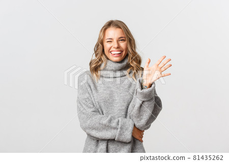 Image of friendly happy woman in grey sweater saying hello, smiling and waving hand to greet person, standing over white background Image of friendly happy woman in grey sweater saying hello, smiling and waving hand to greet person, standing over white background 81435262