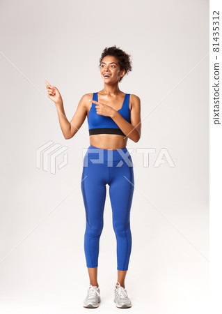 Full length of surprised smiling african-american sportswoman, looking happy at something, pointing fingers left, standing over white background 81435312