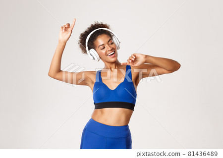 Sport and lifestyle concept. Happy attractive african-american fitness woman dancing, listening music in headphones during workout, white background Sport and lifestyle concept. Happy attractive african-american fitness woman dancing, listening music in headphones during workout, white background 81436489
