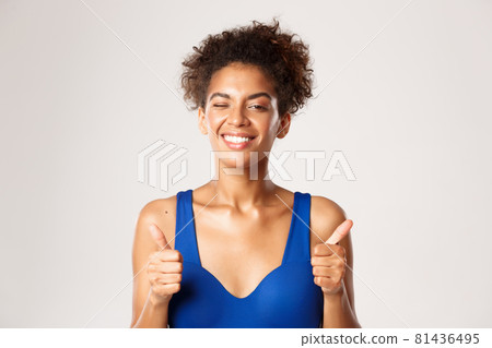 Cheerful african-american female athlete in workout clothing, showing thumbs-up and winking, recommend something about sport, white background Cheerful african-american female athlete in workout clothing, showing thumbs-up and winking, recommend something about sport, white background 81436495