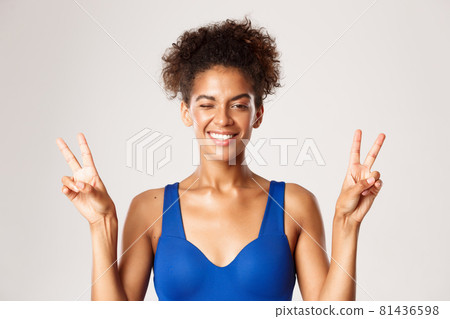 Cheerful african-american fitness girl in blue workout clothing, showing peace signs and smiling happy, standing over white background 81436598