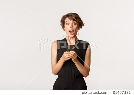 Amazed beautiful female in elegant clothes holding smartphone, open mouth surprised after reading message, standing white background 81437321
