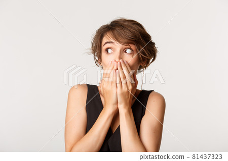 Close-up of shocked gossip girl looking left, cover mouth with hands, standing over white background Close-up of shocked gossip girl looking left, cover mouth with hands, standing over white background 81437323