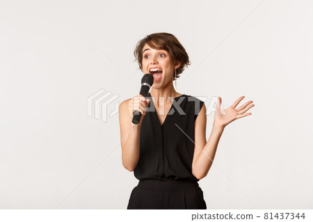 Carefree funny girl singing karaoke, looking left and holding microphone, standing over white background 81437344
