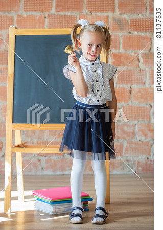 Cute little girl in school uniform posing next to school board with a bell Cute little girl in school uniform posing next to school board with a bell 81437835
