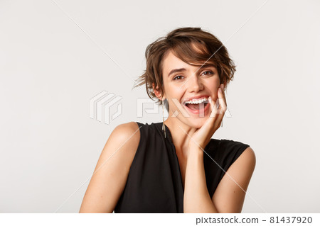 Gorgeous elegant woman laughing flirty and touching face, standing over white background 81437920