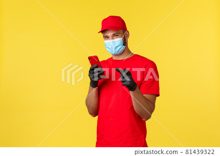 Food delivery, tracking orders, covid-19 and self-quarantine concept. Young courier, employee in medical mask and gloves, using mobile phone and pointing finger camera, yellow background Food delivery, tracking orders, covid-19 and self-quarantine concept. Young courier, employee in medical mask and gloves, using mobile phone and pointing finger camera, yellow background 81439322