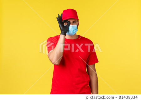 Express delivery during pandemic, covid-19, safe shipping, online shopping concept. Cute courier in medical mask and red uniform over yellow background, show okay sign near eye, guarantee quality Express delivery during pandemic, covid-19, safe shipping, online shopping concept. Cute courier in medical mask and red uniform over yellow background, show okay sign near eye, guarantee quality 81439334