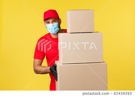 Express delivery during pandemic, covid-19, safe shipping, online shopping concept. Enthusiastic young courier in red uniform, medical mask, standing near piles of boxes with client orders Express delivery during pandemic, covid-19, safe shipping, online shopping concept. Enthusiastic young courier in red uniform, medical mask, standing near piles of boxes with client orders 81439393